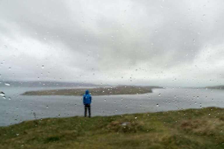 Person in blue jacket stands on the rainy coast of Scotland, overlooking islands.